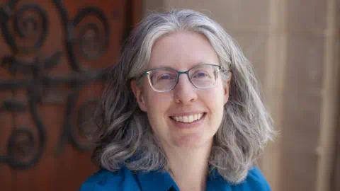 Professor Elizabeth Daniels A woman with grey hair and glasses smiles in front of a grand wooden door